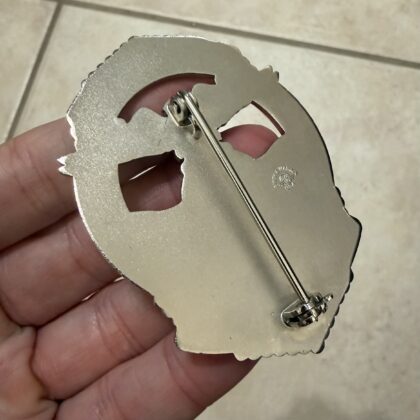 Close-up of a shiny scalloped-edge metal badge being held, showing a pin backing on the back side.